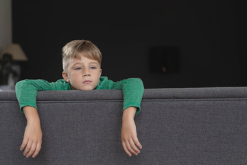 Boy leaning over gray couch in living room, gazing at side table lamp and potted plant