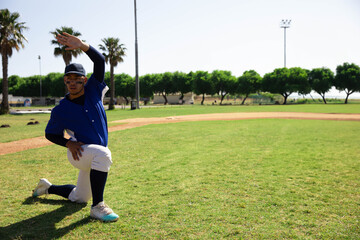 Teenage boy stretching right arm while kneeling on baseball field near dirt infield, copy space