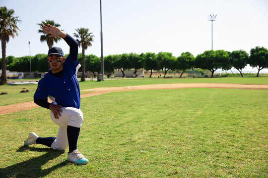 Teenage boy stretching right arm while kneeling on baseball field near dirt infield, copy space - Powered by Adobe