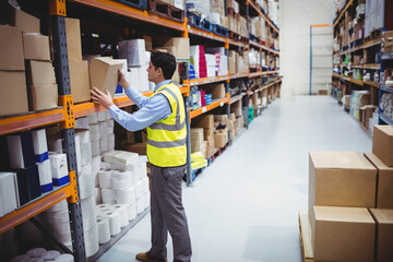 Warehouse worker reaching for cardboard box on tall metal shelf in brightly lit warehouse aisle