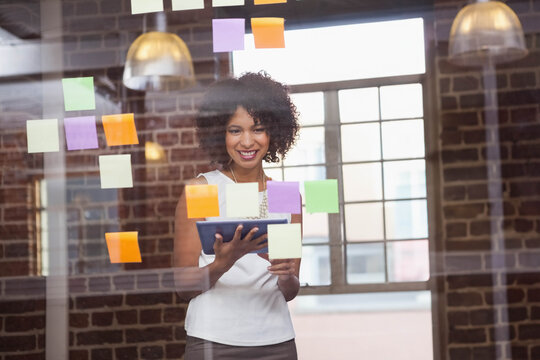 African American woman in business attire using tablet behind board with sticky notes at workspace - Powered by Adobe