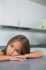 Child girl leaning forward on gray kitchen countertop, gazing into camera beside white cabinets