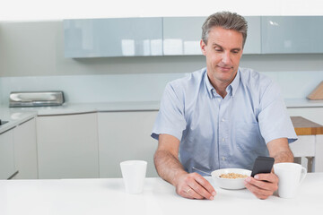 Middle-aged man holding smartphone and eating cereal at kitchen counter near white mugs and toaster