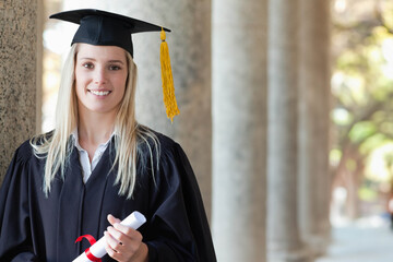 Woman wearing academic gown and mortarboard holding diploma standing at stone colonnade on campus