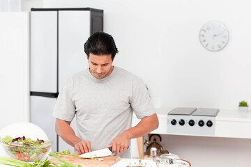 Mid adult male standing in white kitchen slicing celery on wooden cutting board beside glass bowl