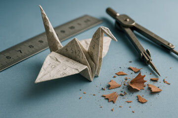Origami paper crane with mathematical formulas surrounded by ruler, compass, and pencil shavings on blue background
