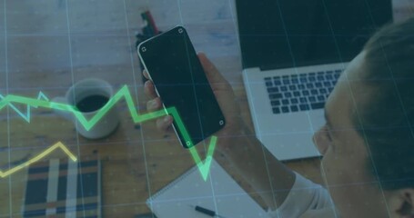 Mature woman picking smartphone and studying while animated financial charts undulating over desk - Powered by Adobe