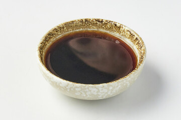 Small ceramic bowl filled with dark brown dipping sauce, commonly used in Japanese cuisine, placed on white background. bowl has textured, rustic design, adding artisanal touch