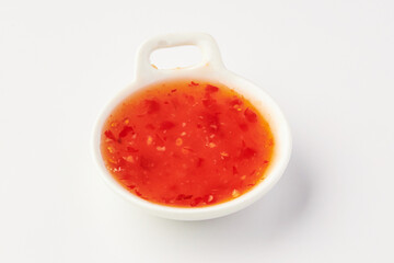 Small white ceramic bowl filled with vibrant red sweet chili sauce, featuring visible chili flakes and seeds, placed on clean white background. sauce appears glossy and appetizing