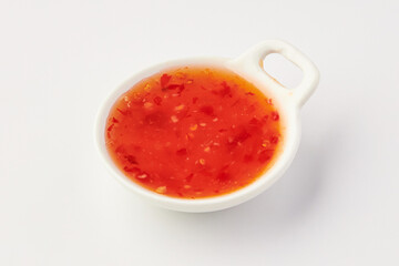 Small white bowl filled with vibrant red sweet chili sauce, showcasing visible chili flakes and seeds, placed on clean white background. sauce appears glossy and appetizing