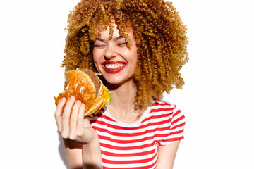 Smiling young woman with curly hair enjoying a delicious hamburger, wearing a striped shirt, on a...