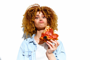 Cheerful young woman with curly hair enjoying a slice of pizza against a bright white background,...