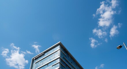 Modern glass building against a vibrant blue sky, accented by fluffy white clouds and a streetlight