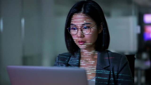 A woman, illuminated by code projected onto her face, looks intently at a laptop in a dimly lit office setting