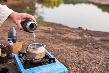 Person pouring water in a kettle to heat it with a travel stove on the bank of a stream, at...