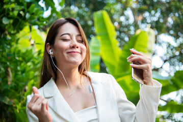 Happy asian woman use headphones listen to music from smartphone outdoor in green park nature....