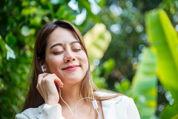 Happy asian woman use headphones listen to music from smartphone outdoor in green park nature....