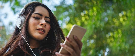 Banner Happy asian woman use headphones listen to music from smartphone outdoor in green park....