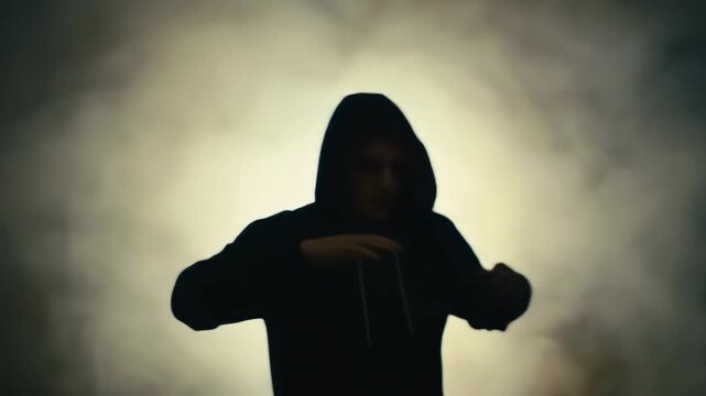 Mysterious silhouette of a person in a hoodie gesturing powerfully against a smoky, backlit background.