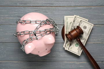 Piggy bank tied in chain and judge's gavel with dollar banknotes on grey wooden background. Debt...