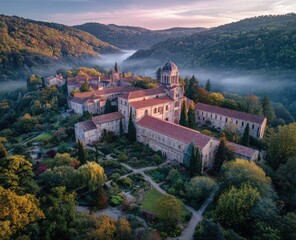 Obraz premium Serene abbey nestled in a misty valley at sunrise, surrounded by autumnal foliage and extensive gardens