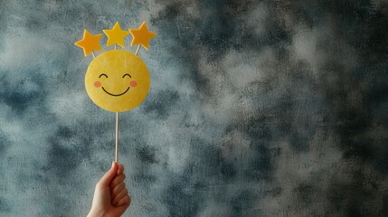 A cheerful hand holding a yellow smiley face on a stick, featuring three stars. The background is a textured blue-gray hue, creating a playful and vibrant mood.