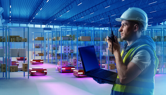 Technician in safety gear with laptop and radio supervises autonomous robots in a smart warehouse with automated storage. Focus on logistics and robotics. Industry: Warehouse, Logistics. - Powered by Adobe