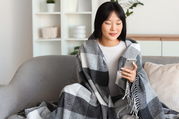 Young Asian woman with soft plaid using mobile phone on sofa at home