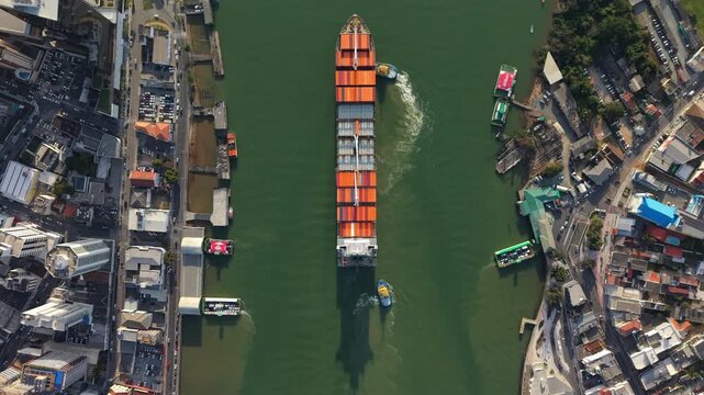 Container ship moves through the Itaja&iacute; Port channel in Santa Catarina, Brazil, guided by tugboats between urban docks and industrial areas, captured from above in a precise top-down slow drone shot.