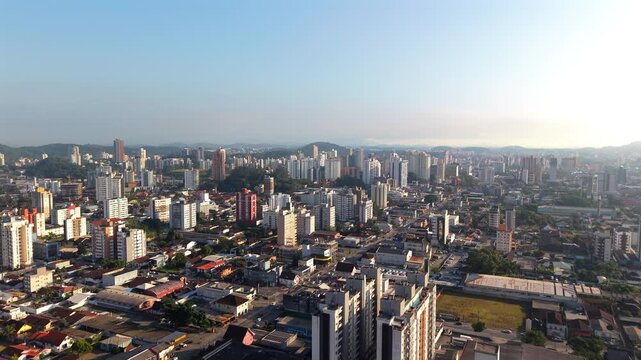 Drone glides over Joinville, Santa Catarina, Brazil, revealing a dense urban grid, notable buildings like Joinville Cathedral, shopping centers, parks, and Serra do Mar mountains in the distance.