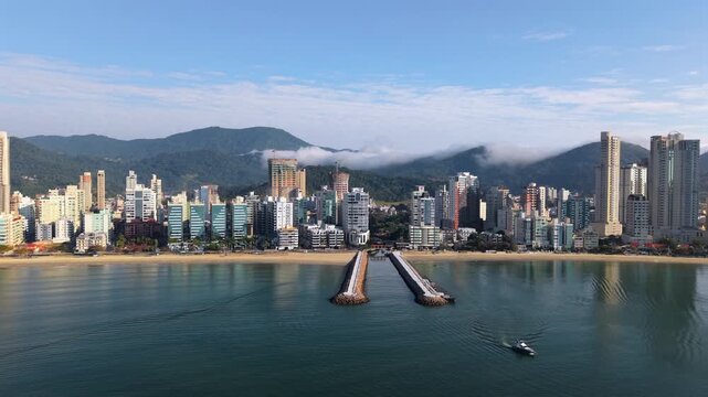 Slow drone pans Itapema Meia Praia skyline and beachfront in Paran&aacute;, Brazil, showcasing sandy beach, oceanfront high-rises, marina piers, and the Serra do Mar mountains under a partly cloudy blue sky.
