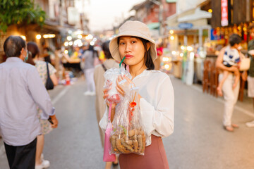 Young Asian woman holding a bag of snacks and sipping a beverage from a straw while exploring a vibrant street market at sunset