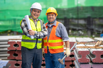 Two construction workers wea safety helmets and reflective vests standing outdoors at a construction site with bricks and safety equipment, discussing project plans