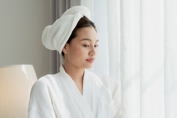 Asian teenage girl in white bathrobe with towel on hair looking through window in calm natural...