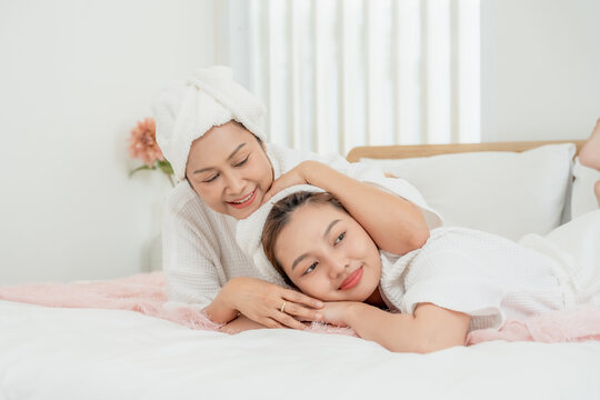 Asian senior mother hugging teenage daughter lying together on bed during home spa wearing white robes and towels on heads expressing family love warmth comfort in skincare routine