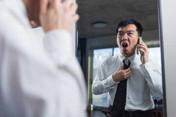 A young man holds a phone to his ear and yells while adjusting his tie in front of a mirror. The...