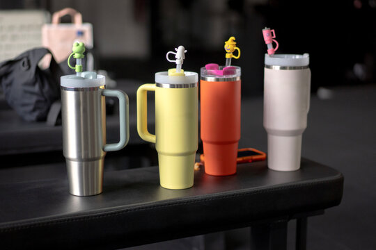 Four colorful insulated tumblers or travel mugs with cute straw toppers lined up on a bench in a gym. Concept of hydration, fitness, and lifestyle.