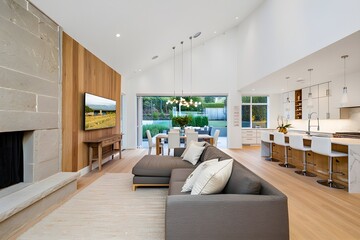 Obraz premium Modern open-plan living area with gray sectional sofa, light wood floors, stone fireplace, and kitchen island with bar stools, overlooking verdant backyard through expansive folding glass doors.