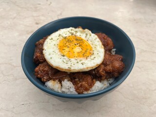 Close-up of rice bowl topped with fried chicken and sunny side up egg, garnished with herbs. Asian comfort food served in blue bowl on marble background.