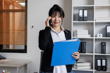 A cheerful, beautiful Young Asian businesswoman is talking on the phone with her client while...