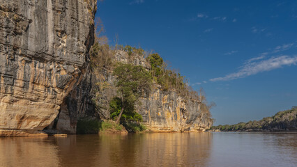 Fototapeta premium A calm red-brown African river. There is green vegetation on the steep limestone shores. Reflection on the water. Blue sky, clouds. Madagascar. Manambolo river.