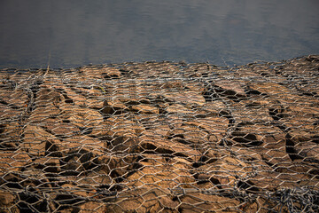 Close-up of gabion retaining the shoreline by a lake, featuring interwoven rocks and wire, ideal for erosion control and natural landscape themes.