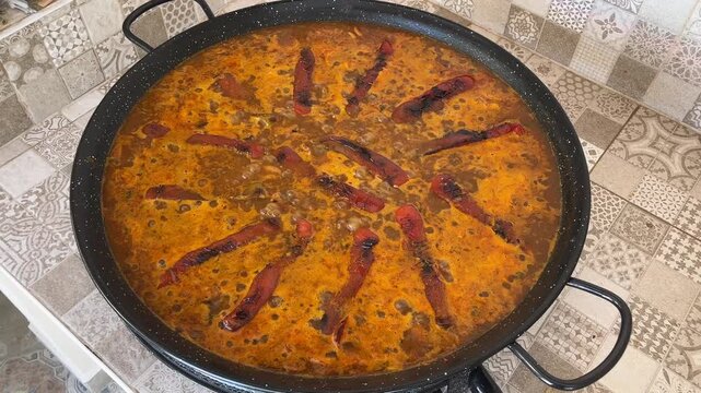 Large Fideu&agrave; pan cooking on a patterned tile countertop, with roasted red peppers arranged in a sunburst pattern, during summer vacations in Gandia, Valencia, Spain