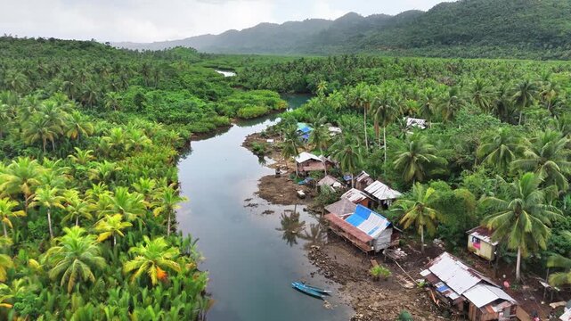 A tranquil river winds through dense mangroves and palm forests beside a remote village in Mataob Maasin, Siargao Islands, Philippines, nestled against green hills under a cloudy sky