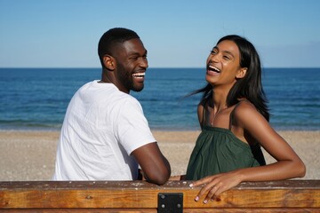 Happy diverse couple laughing on beach bench enjoying summer vacation together getaway