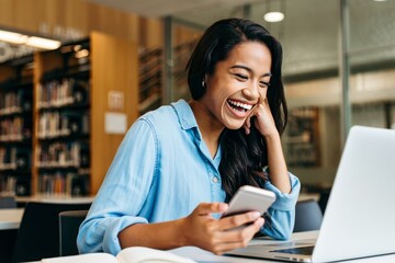 Young woman laughing looking at smartphone in library with laptop and books study