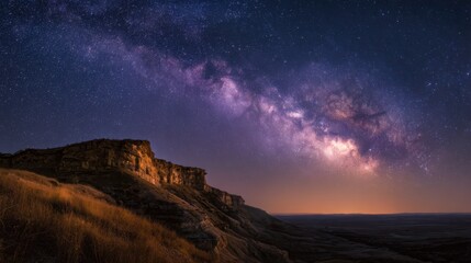 A breathtaking night sky showcases the Milky Way galaxy above a rugged cliff with dry grass, blending cosmic beauty with earthly wilderness under a star-studded horizon.