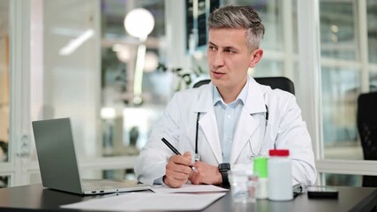 Experienced confident male doctor wearing a white lab coat and stethoscope thoughtfully writes notes at a desk in a modern clinic office, looking focused and dedicated. - Powered by Adobe