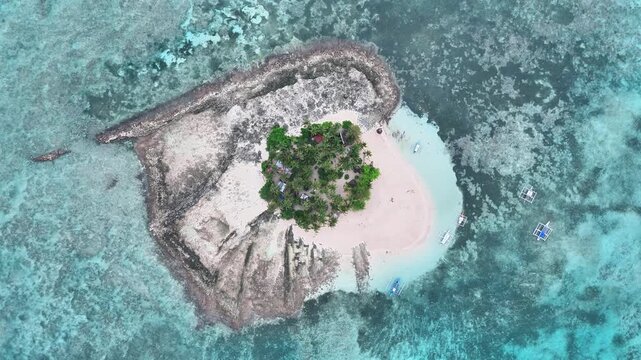 Aerial view of Guyam Island in Siargao, Philippines, featuring a circular sandy beach, lush palm trees, turquoise shallows, coral reef surroundings, and small boats approaching from the vibrant sea