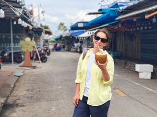 A woman in sunglasses and a yellow shirt holds a fresh coconut with a straw